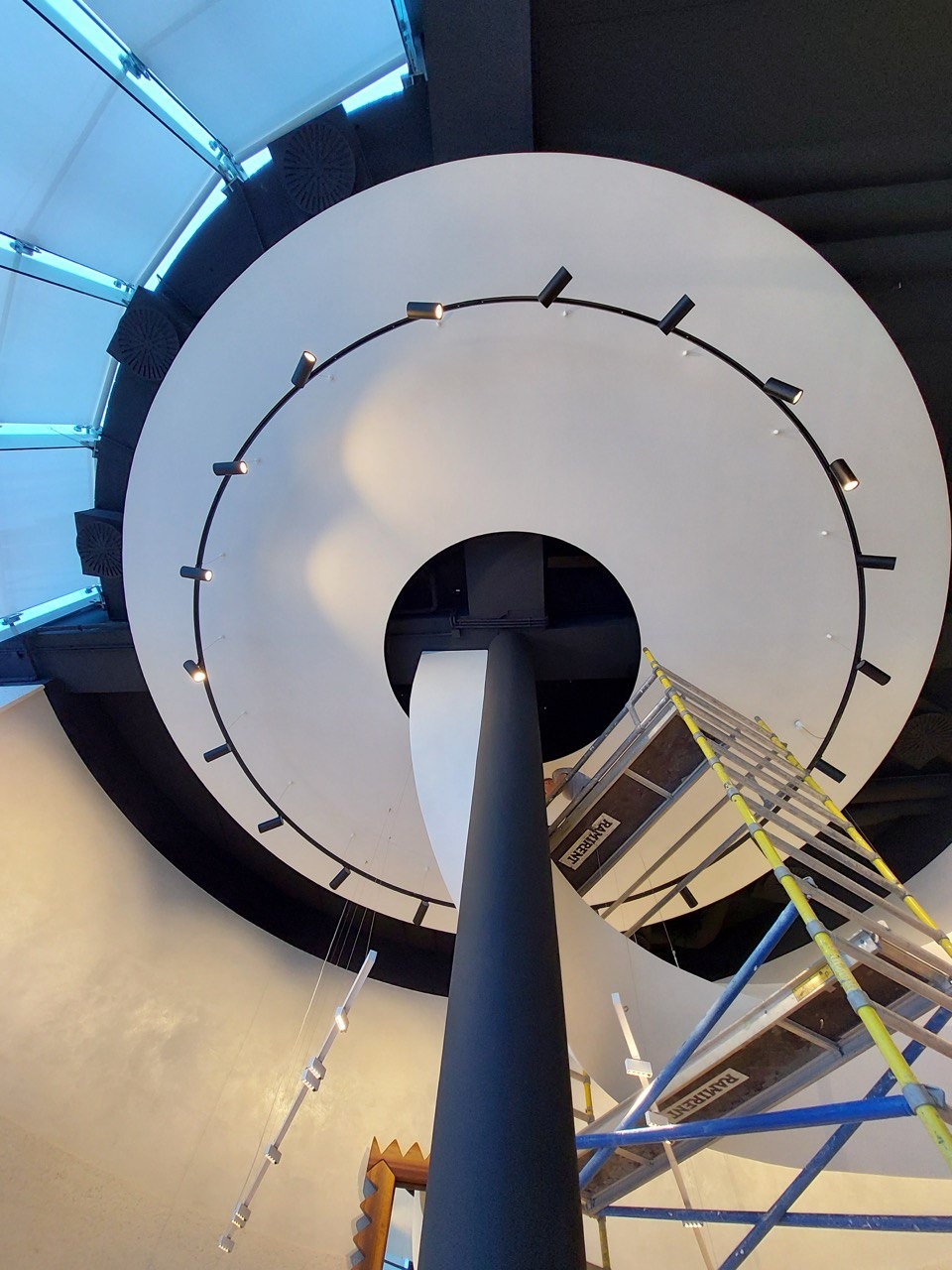 Okrągła konstrukcja sufitowa z zamontowanymi reflektorami punktowymi, widok z dołu, rusztowanie w tle sugerujące prace montażowe.