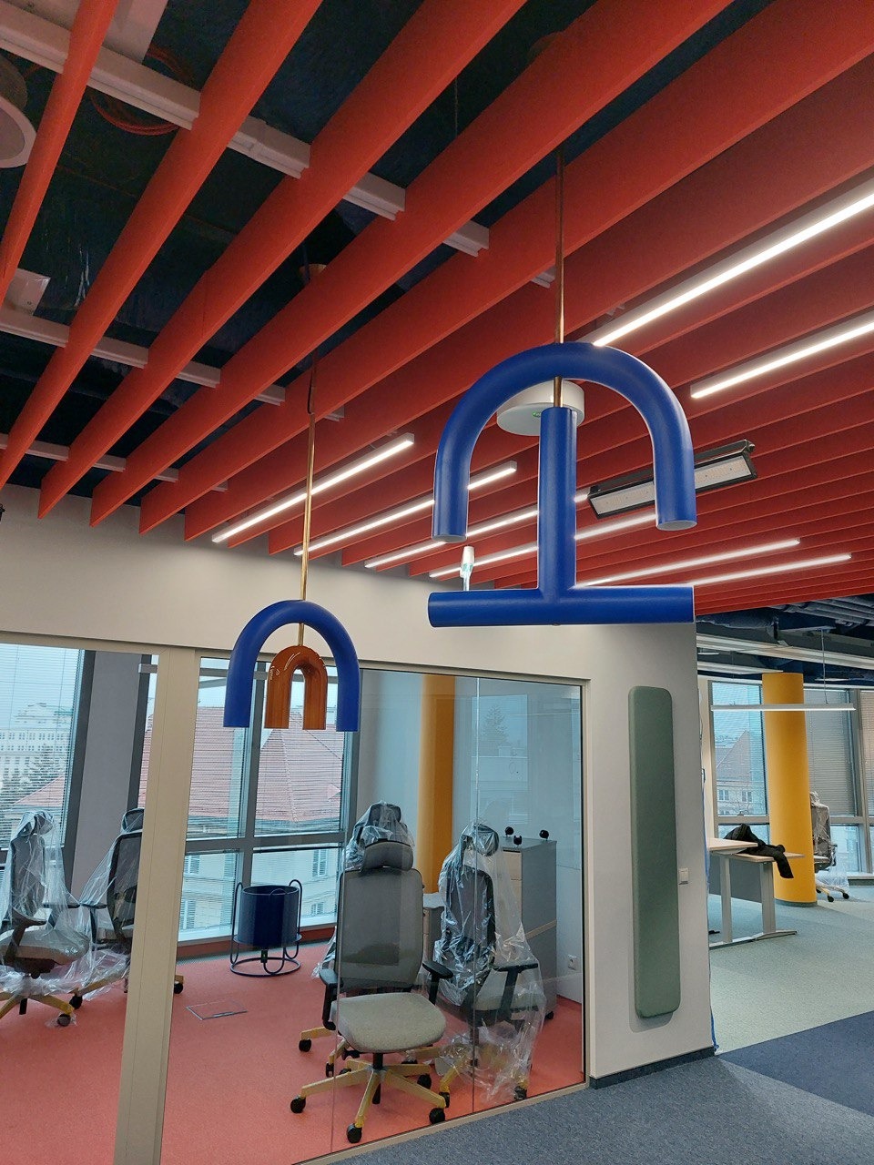 Biuro z nowoczesnym oświetleniem sufitowym w postaci kolorowych, geometrycznych lamp wiszących i listew LED, widok przez szklaną ścianę na pomieszczenie z meblami biurowymi w folii ochronnej.