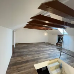 Poddasze w Białymstoku po renowacji, z drewnianą podłogą, białymi ścianami, drewnianymi belkami stropowymi i schodami w trakcie wykończenia, widoczna drabina i butelka wody.