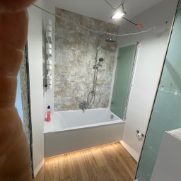 Przedsiębiorstwo Inwestycyjne KBK Dawid Kubik - Wykończona łazienka z wanną, prysznicem i drewnopodobną podłogą. Częściowo widoczna płyta karton gips. Ujęcie z palcem w kadrze.