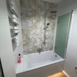 Przedsiębiorstwo Inwestycyjne KBK Dawid Kubik - Nowoczesna łazienka po remoncie: biała wanna z szarą mozaiką, szklany parawan, grzejnik drabinkowy i drewnopodobna podłoga z subtelnym oświetleniem LED.