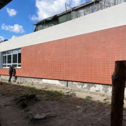 Przedsiębiorstwo Inwestycyjne KBK Dawid Kubik - Elewacja budynku wykończona ceglastymi płytkami, widoczne okna i fragment terenu. Dwóch mężczyzn przy oknie. Częściowo widoczne rusztowanie.