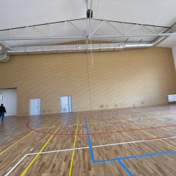 Przedsiębiorstwo Inwestycyjne KBK Dawid Kubik - Sala gimnastyczna po remoncie: parkiet z liniami boiska, ceglana ściana, wentylacja pod sufitem, osoba w oddali. Widok na przestronne, jasne wnętrze.