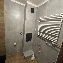 Przedsiębiorstwo Inwestycyjne KBK Dawid Kubik - Wykończona łazienka z szarymi płytkami, drewnopodobną podłogą i białym grzejnikiem drabinkowym. Widoczne elementy instalacji i wentylacji.