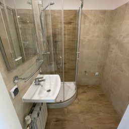 Przedsiębiorstwo Inwestycyjne KBK Dawid Kubik - Jasna łazienka po remoncie: kabina prysznicowa z zaokrąglonym szkłem, umywalka z baterią, grzejnik i beżowe płytki imitujące drewno na ścianach i podłodze.