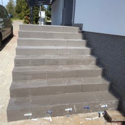 Przedsiębiorstwo Inwestycyjne KBK Dawid Kubik - Wykończone płytkami schody zewnętrzne z widocznymi krzyżykami dystansowymi, świeżo po montażu. Szare płytki na stopniach i cokoliku przy ścianie budynku.