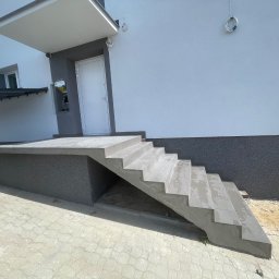 Przedsiębiorstwo Inwestycyjne KBK Dawid Kubik - Wykończone płytkami schody wejściowe do budynku z szarą elewacją i białymi drzwiami. Widoczny fragment chodnika z kostki brukowej i zaparkowany samochód.