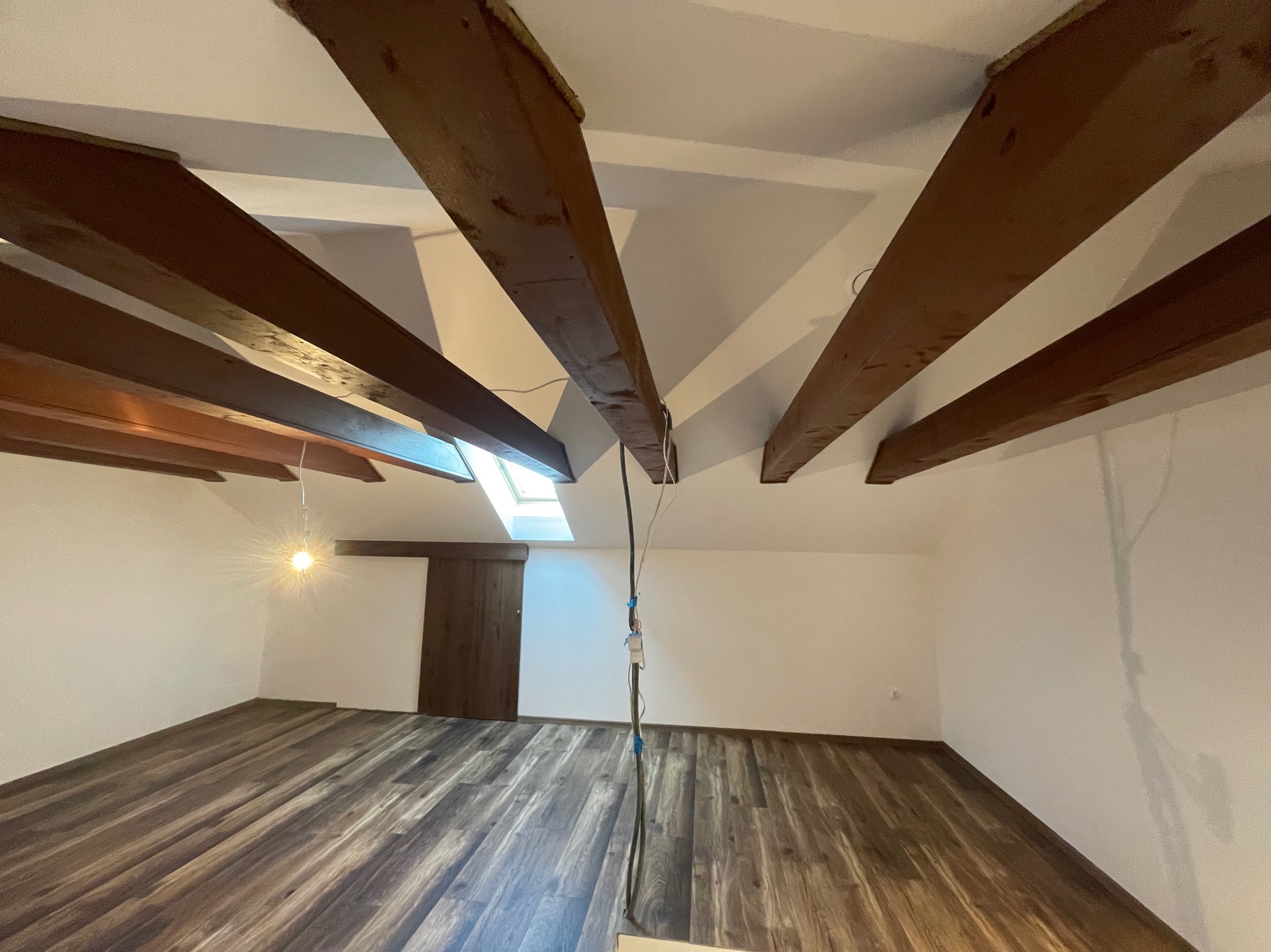 Poddasze po remoncie z drewnianą podłogą, białymi ścianami i odsłoniętymi, ciemnymi belkami stropowymi; widoczne okno dachowe i wisząca żarówka; przewody elektryczne wystające z sufitu.