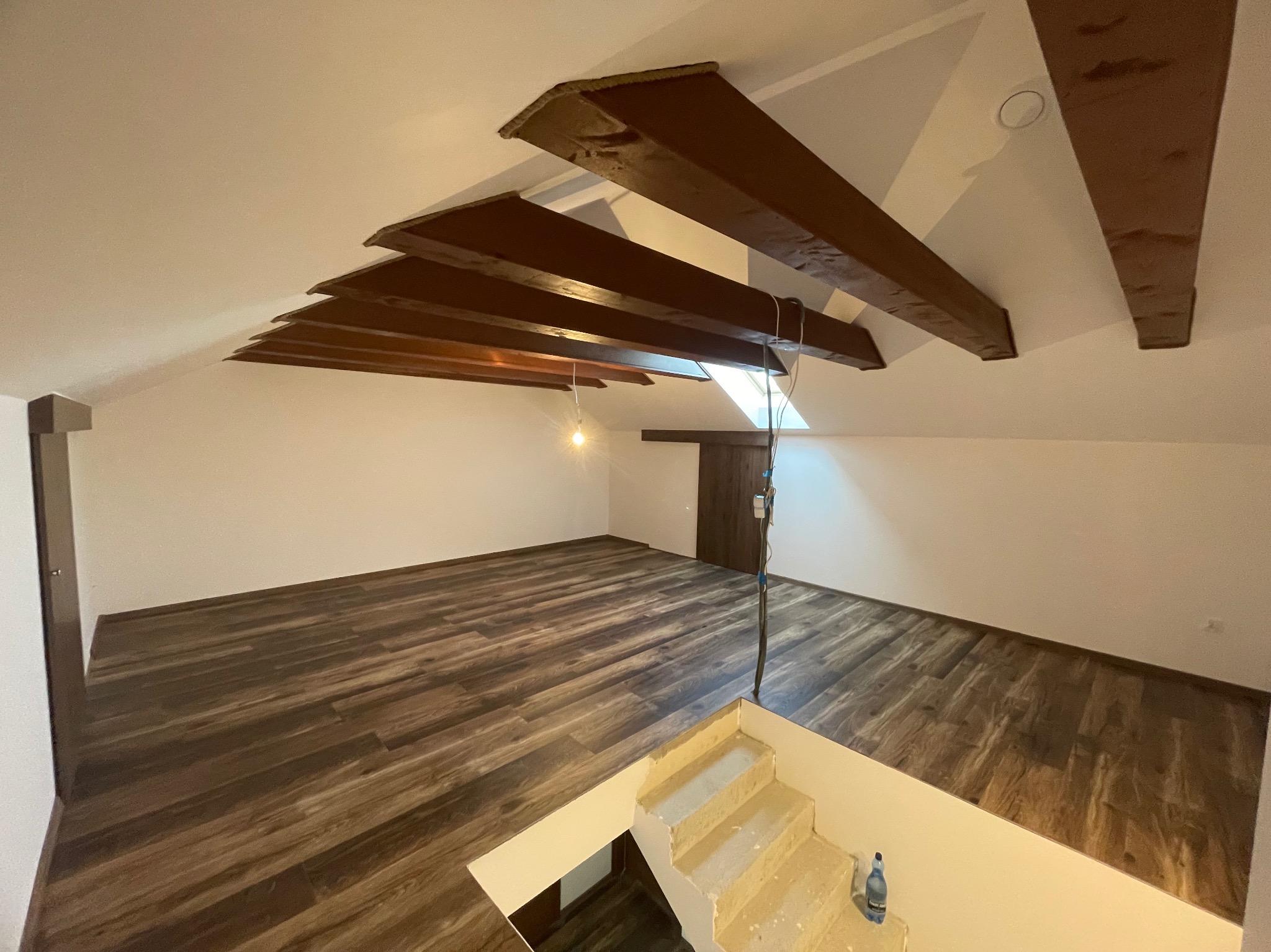 Poddasze po remoncie z drewnianymi belkami stropowymi, wykończone panelami podłogowymi imitującymi drewno; częściowo wykończone schody z butelką wody mineralnej na dole