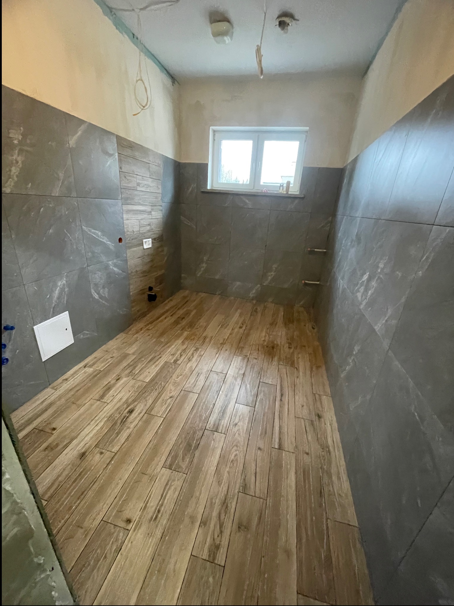 Wnętrze łazienki w trakcie prac wykończeniowych; szare płytki imitujące kamień na ścianach do połowy wysokości, drewnopodobne płytki na podłodze, widoczne wystające kable i rury, surowy stan górnej...