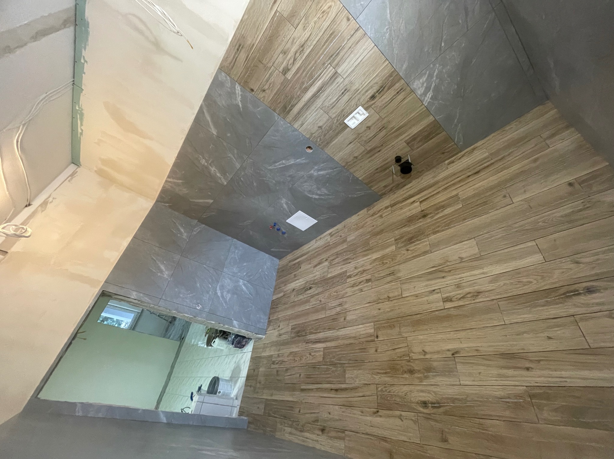 Wnętrze łazienki w trakcie prac wykończeniowych, widoczne ułożone płytki drewnopodobne i szare, surowa ściana z instalacją elektryczną, otwór na okno.