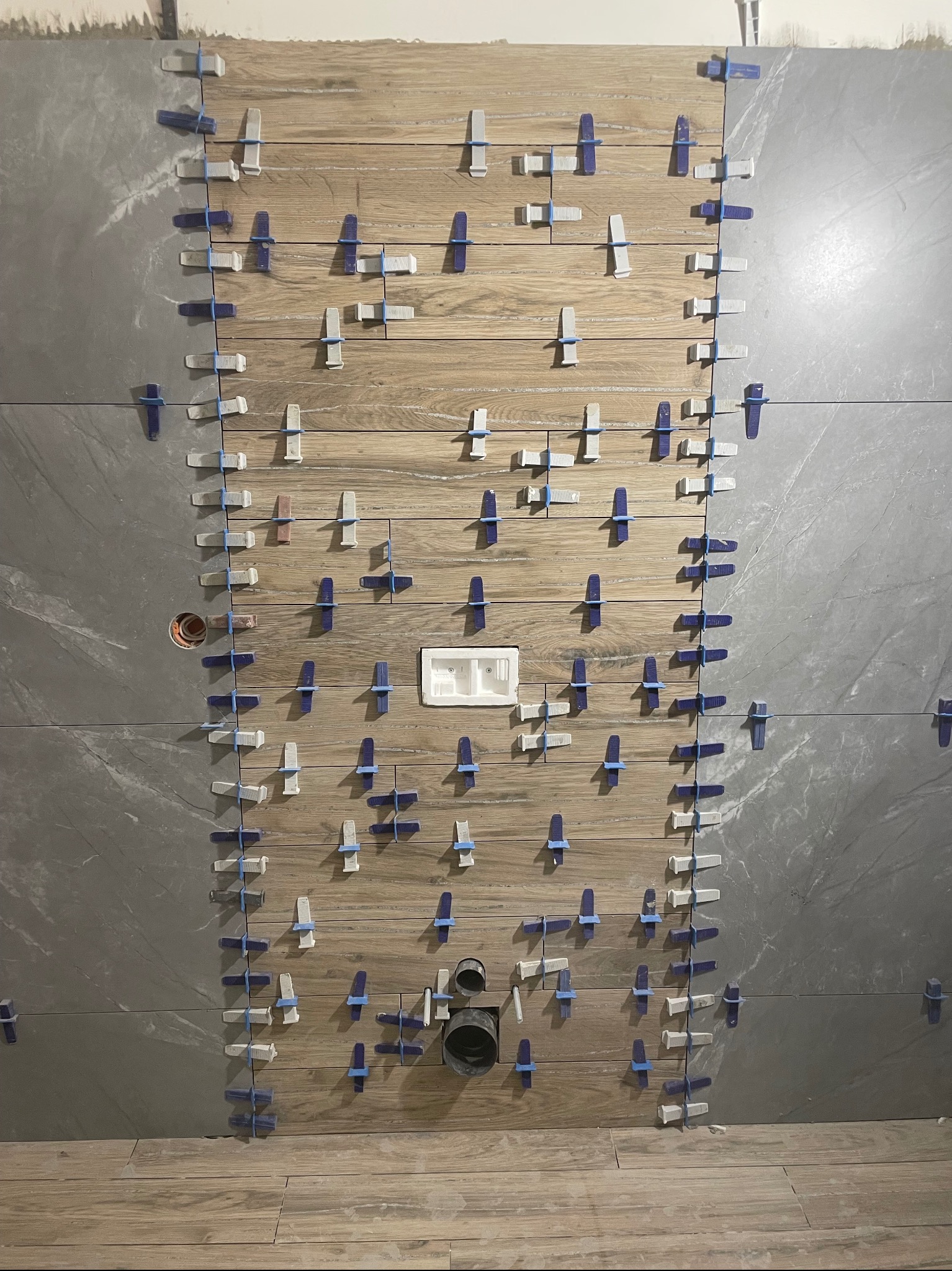 Ściana w trakcie układania płytek, widoczne płytki imitujące drewno oraz szare płytki, użyte klipsy poziomujące, otwór na rurę kanalizacyjną i puszkę elektryczną.