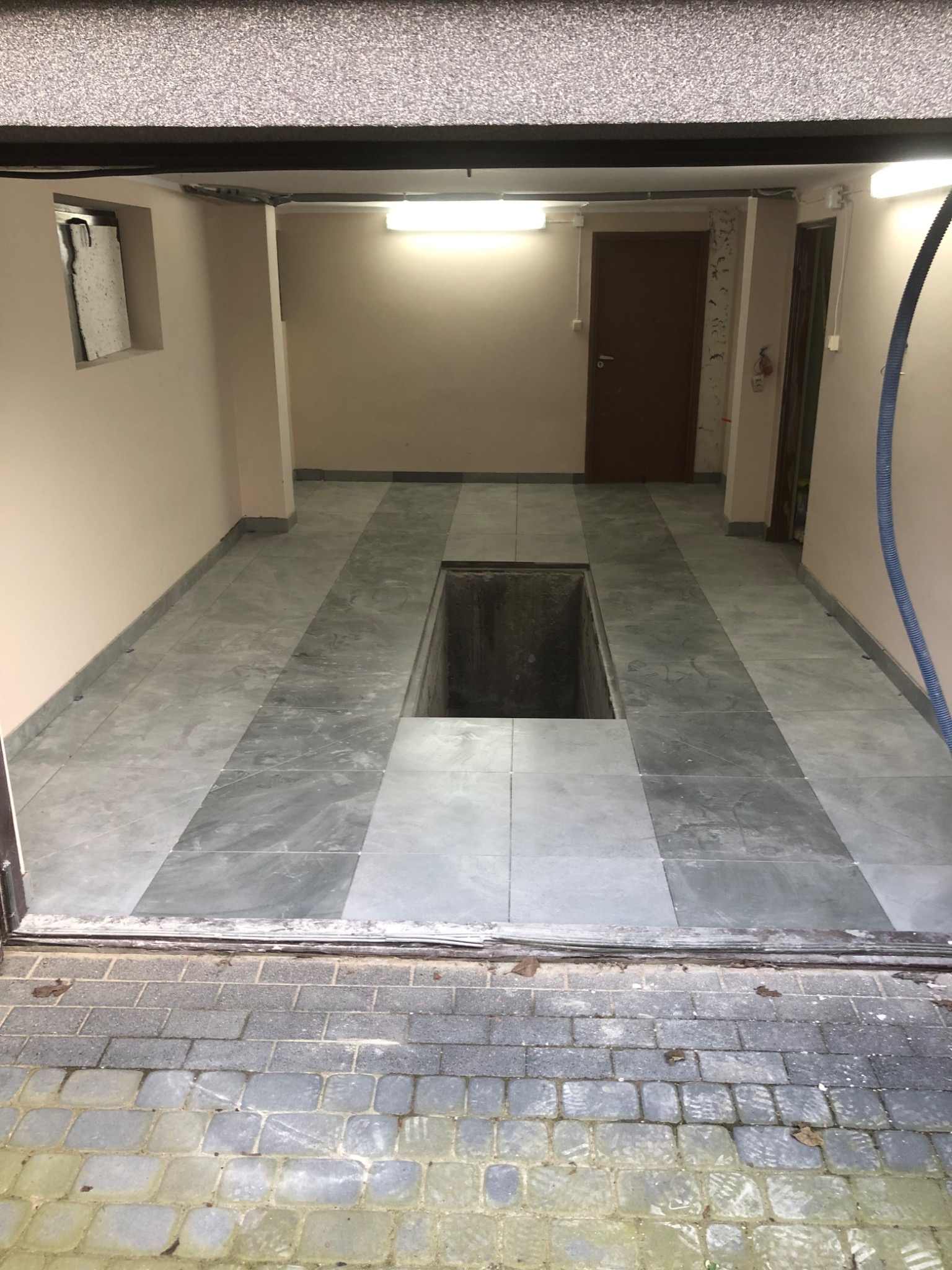 Wykończona podłoga w garażu z szarych płytek, widoczny otwór w podłodze. Na ścianach jasny beż, w tle brązowe drzwi. Widok z zewnątrz, z kostki brukowej.