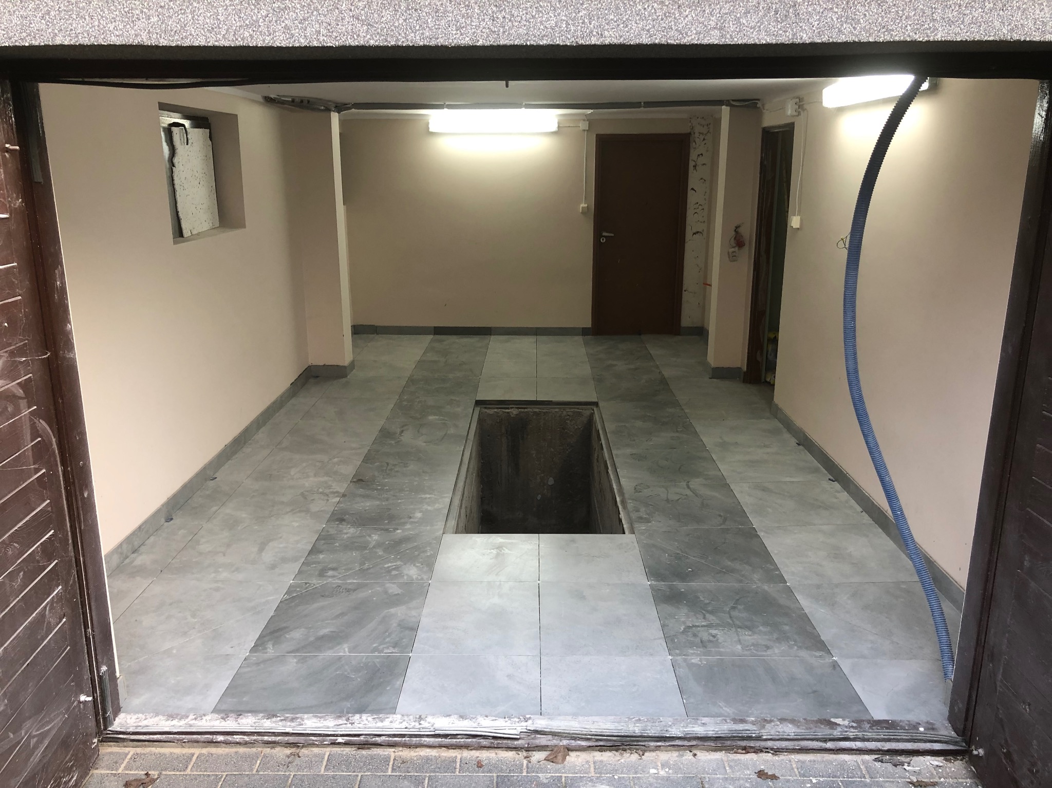 Wykończone płytkami podłogowymi pomieszczenie z otworem w podłodze, widoczne drzwi i rura wentylacyjna. Szare płytki ułożone w pasy.