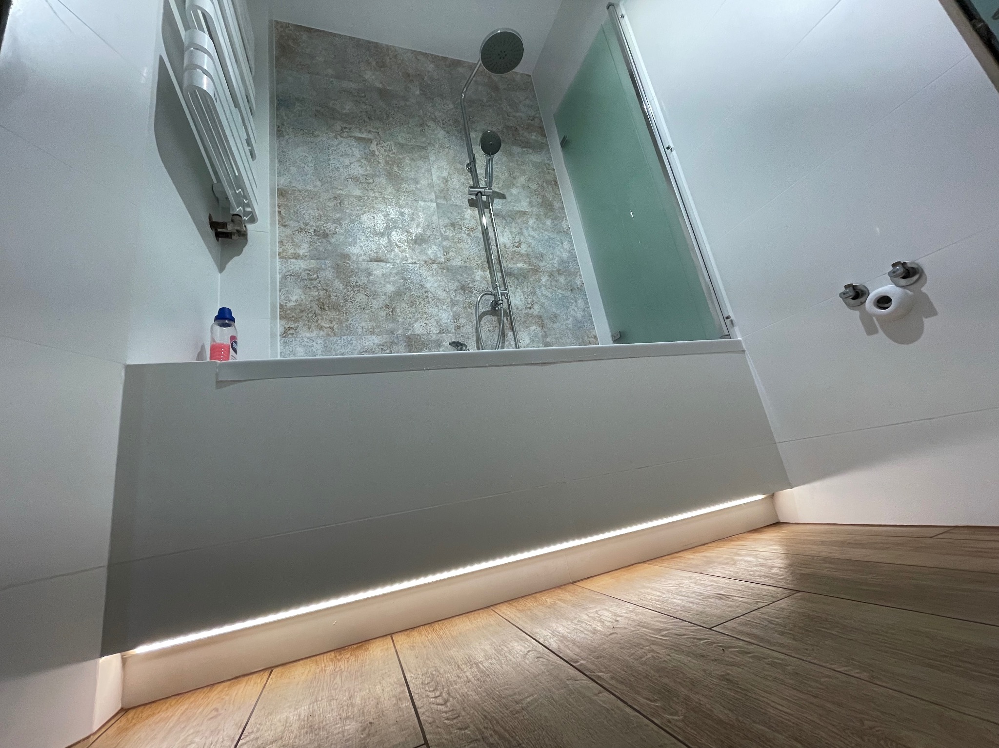 Nowoczesna łazienka po remoncie: kabina prysznicowa, płytki imitujące kamień, podświetlenie LED przy podłodze i drewnopodobne płytki podłogowe.