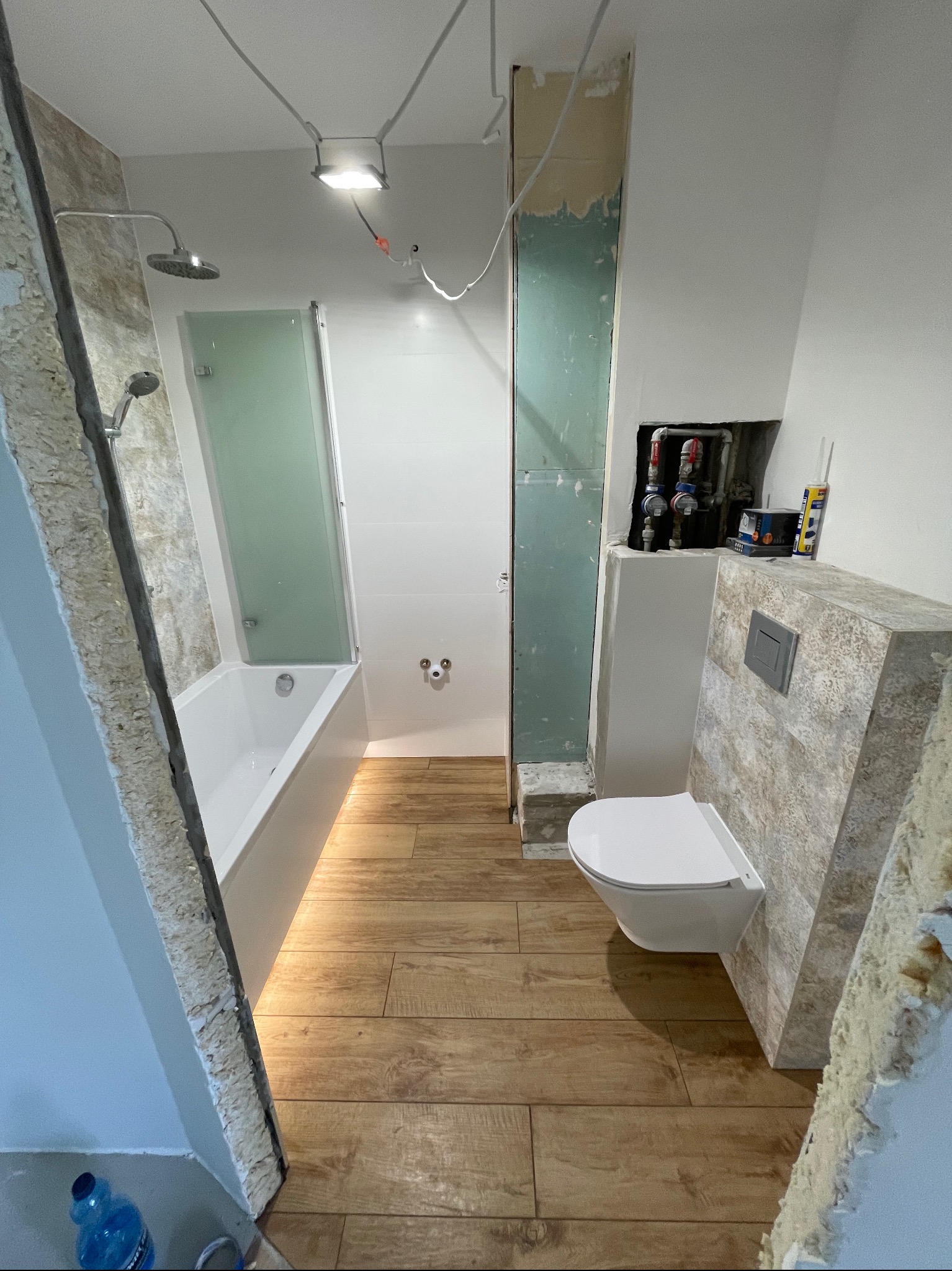Remont łazienki w trakcie: biała wanna z kabiną, toaleta podtynkowa z szarą płytką imitującą kamień, drewnopodobna podłoga, widoczne instalacje i niedokończone ściany.