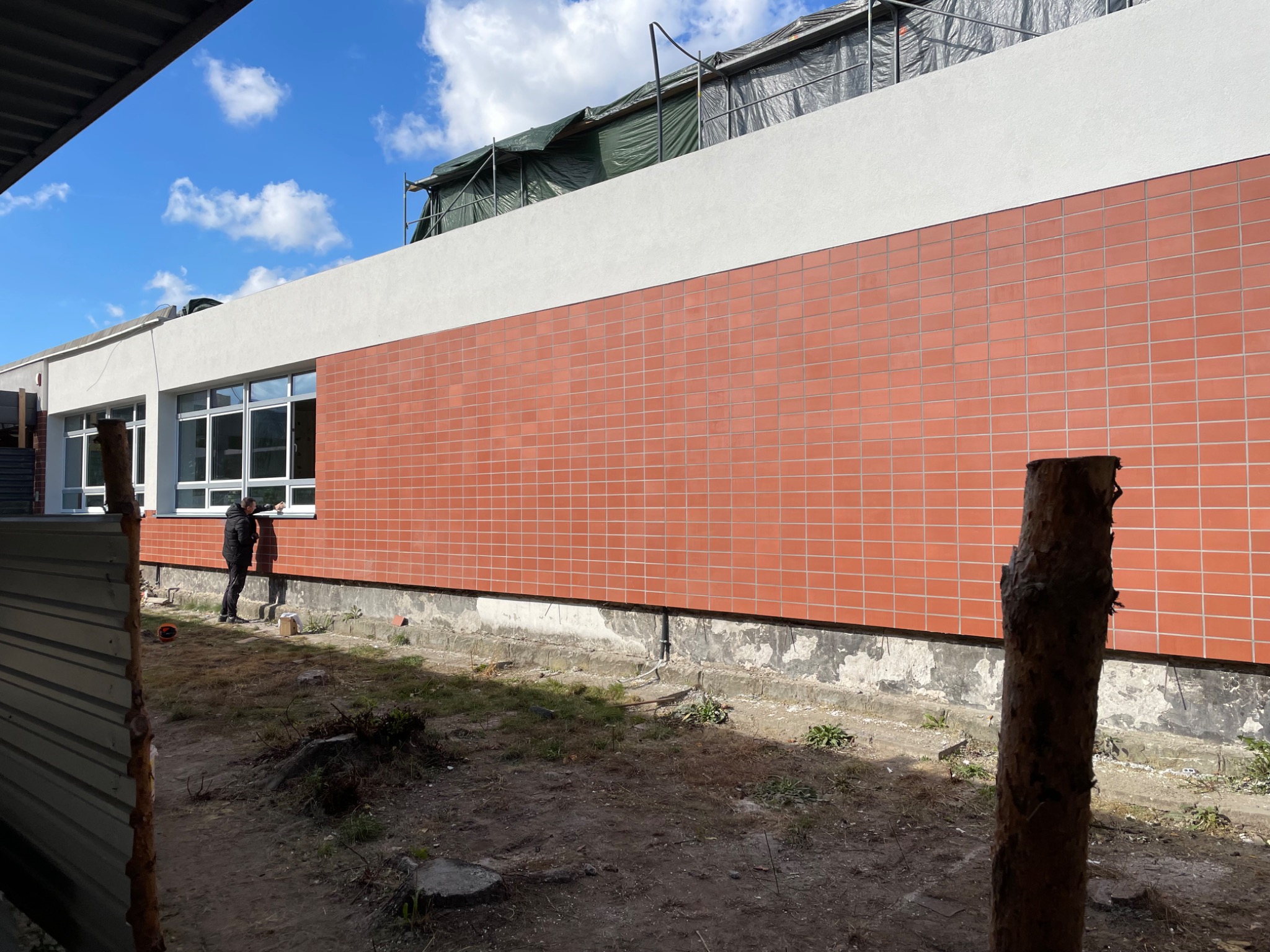 Elewacja budynku wykończona ceglastymi płytkami, widoczne okna i fragment terenu. Dwóch mężczyzn przy oknie. Częściowo widoczne rusztowanie.