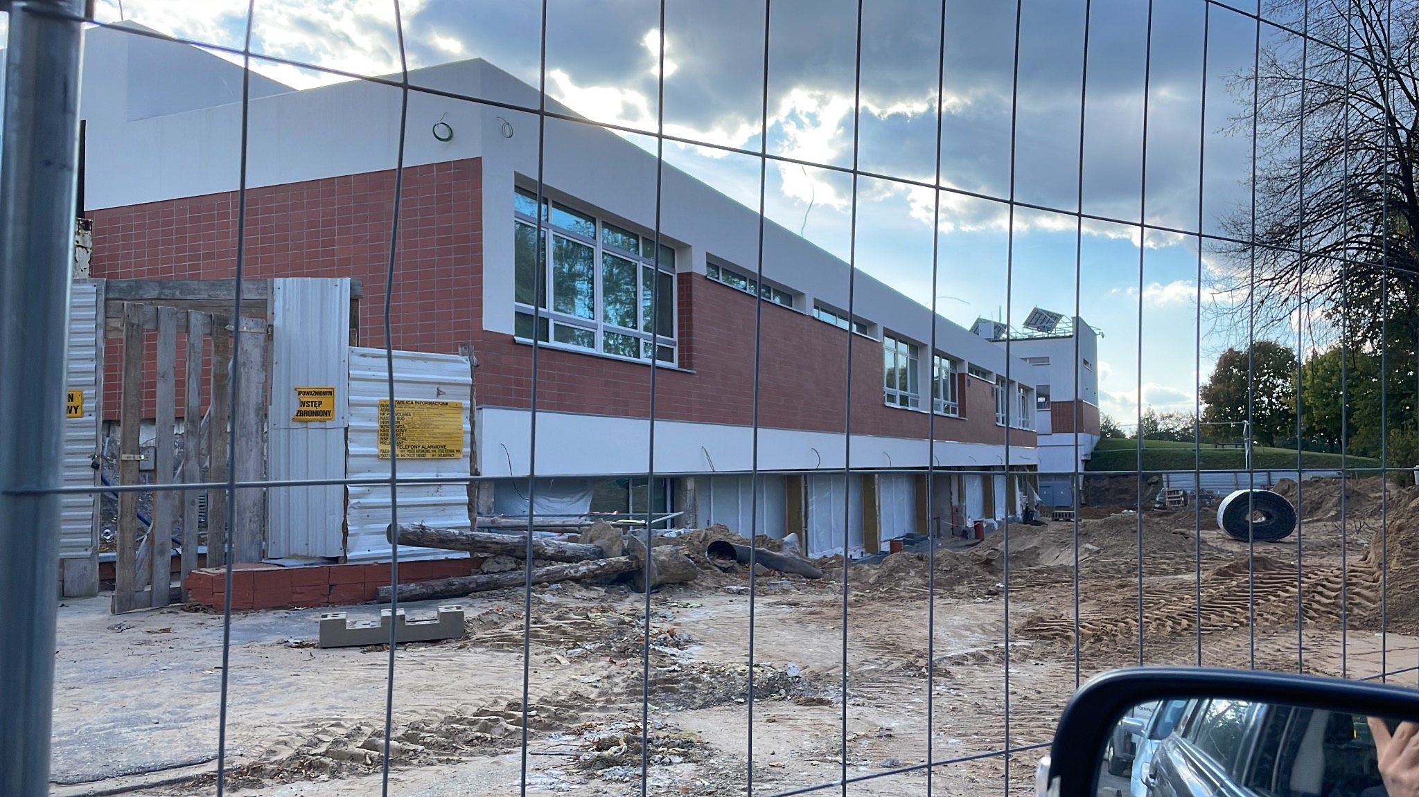 Budynek w trakcie remontu w Białymstoku, widoczny przez ogrodzenie. Prace ziemne, odsłonięte fundamenty, ceglana elewacja i białe elementy wykończeniowe.