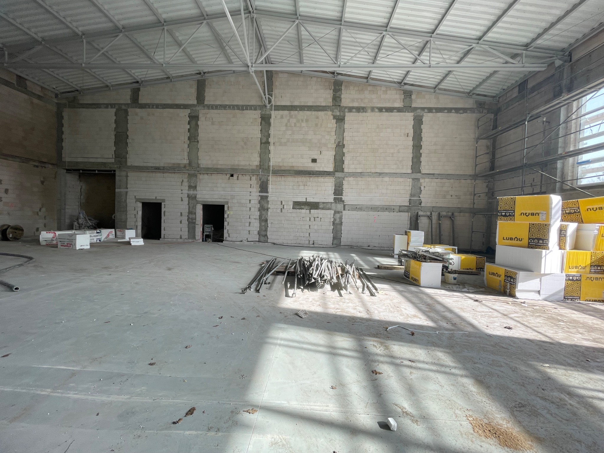 Wnętrze hali w trakcie budowy: surowe ściany z betonu komórkowego, metalowa konstrukcja dachu, materiały budowlane i narzędzia na posadzce. Widoczne ślady prac budowlanych.