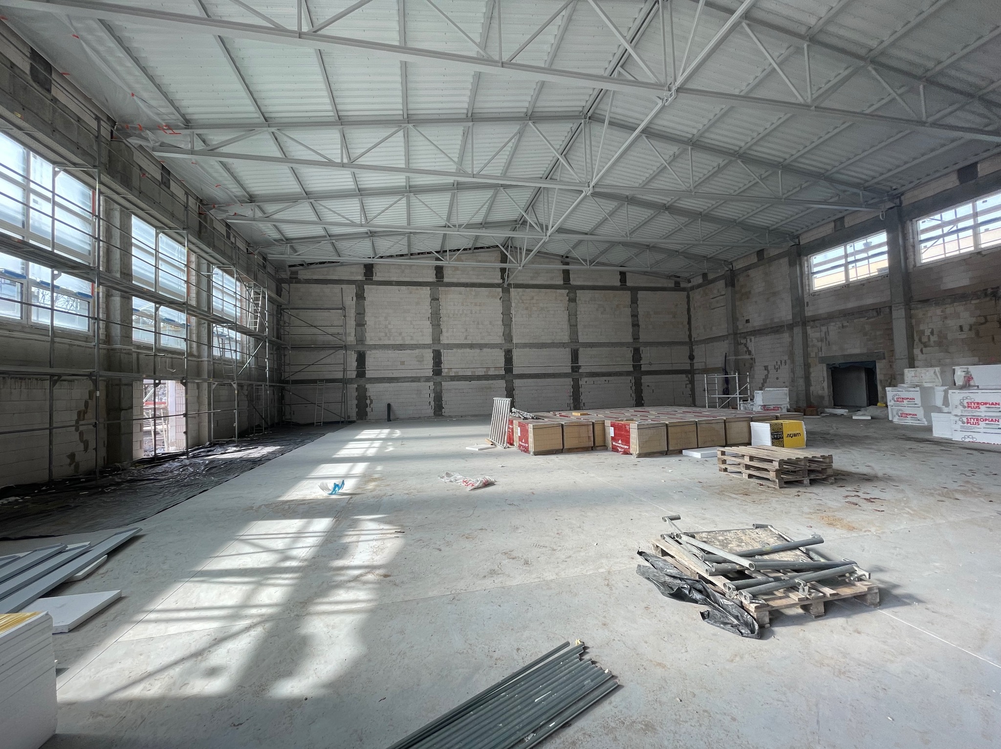 Wnętrze hali w trakcie budowy: surowy beton, rusztowania przy oknach, materiały budowlane na paletach, widoczna konstrukcja dachu. Hala w stanie deweloperskim.