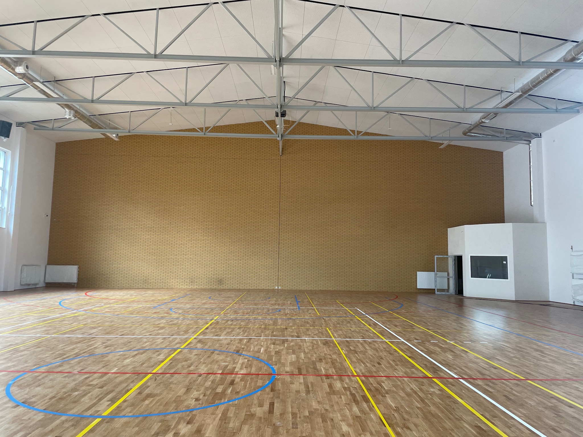 Sala gimnastyczna z parkietem w linie, ceglaną ścianą i metalową konstrukcją dachu. Widoczne okno i biała zabudowa z drzwiami. Oświetlenie górne.