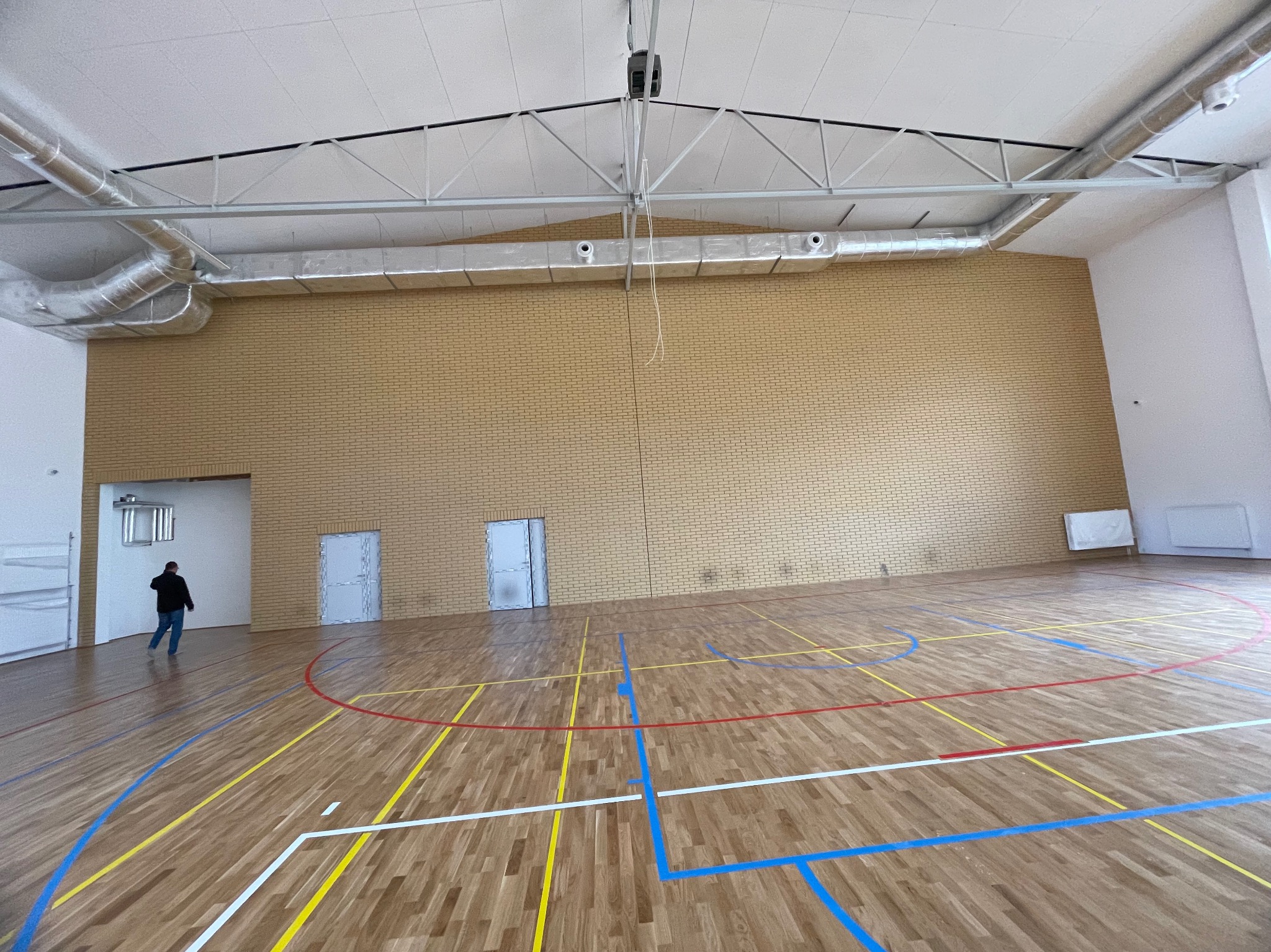 Sala gimnastyczna po remoncie: parkiet z liniami boiska, ceglana ściana, wentylacja pod sufitem, osoba w oddali. Widok na przestronne, jasne wnętrze.