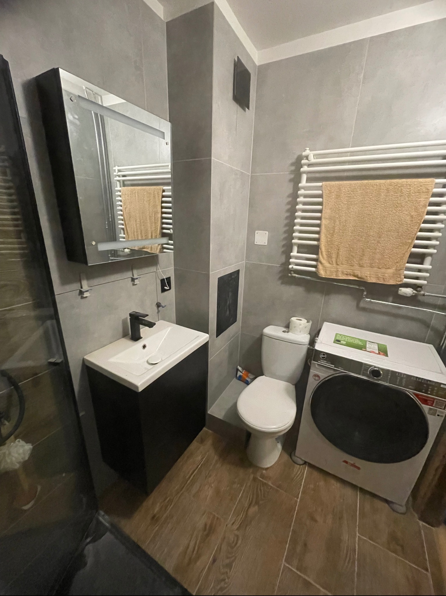 Szara łazienka z drewnopodobną podłogą, umywalką z czarną szafką, toaletą, pralką i grzejnikiem drabinkowym z ręcznikiem. Minimalistyczny design.