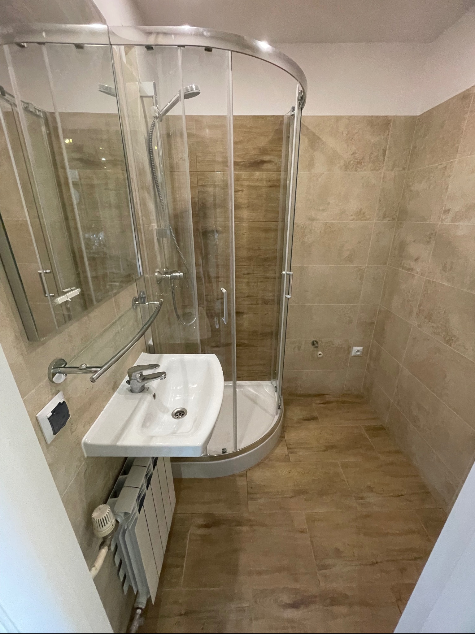 Jasna łazienka po remoncie: kabina prysznicowa z zaokrąglonym szkłem, umywalka z baterią, grzejnik i beżowe płytki imitujące drewno na ścianach i podłodze.