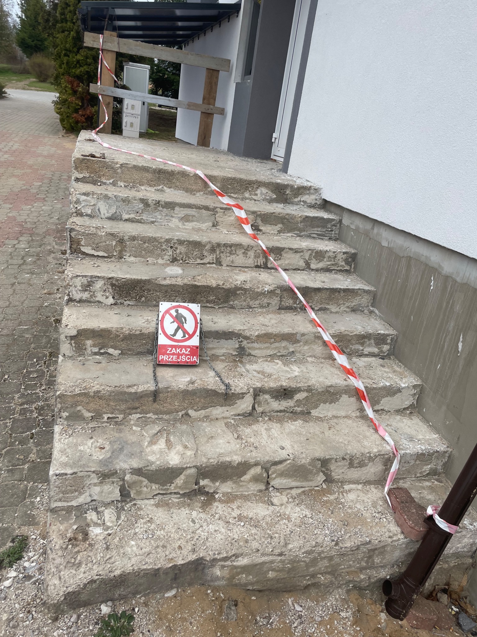 Zniszczone, betonowe schody wejściowe do budynku, zabezpieczone taśmą ostrzegawczą i znakiem zakazu przejścia. Widoczne ślady uszkodzeń i przygotowania do remontu.