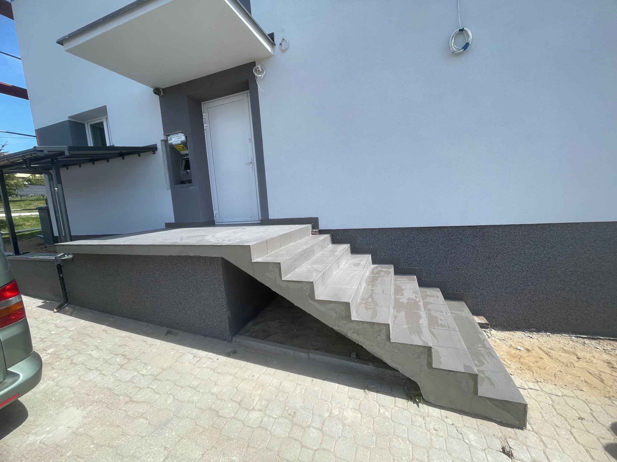 Wykończone płytkami schody wejściowe do budynku z szarą elewacją i białymi drzwiami. Widoczny fragment chodnika z kostki brukowej i zaparkowany samochód.