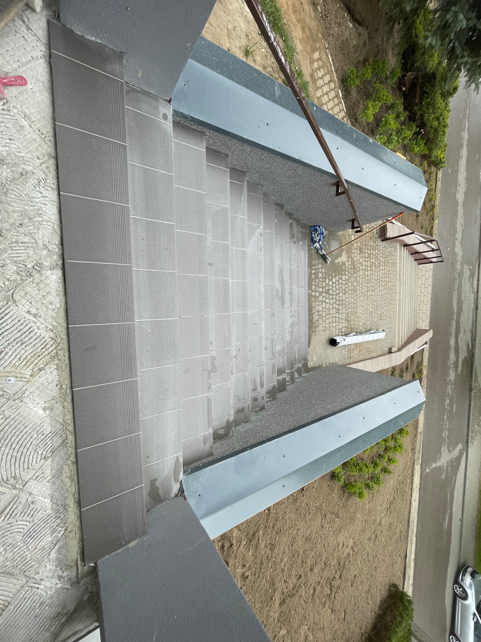 Ułożone szare płytki na schodach zewnętrznych budynku, widok z góry. Częściowo widoczne narzędzia glazurnicze i otoczenie budynku.