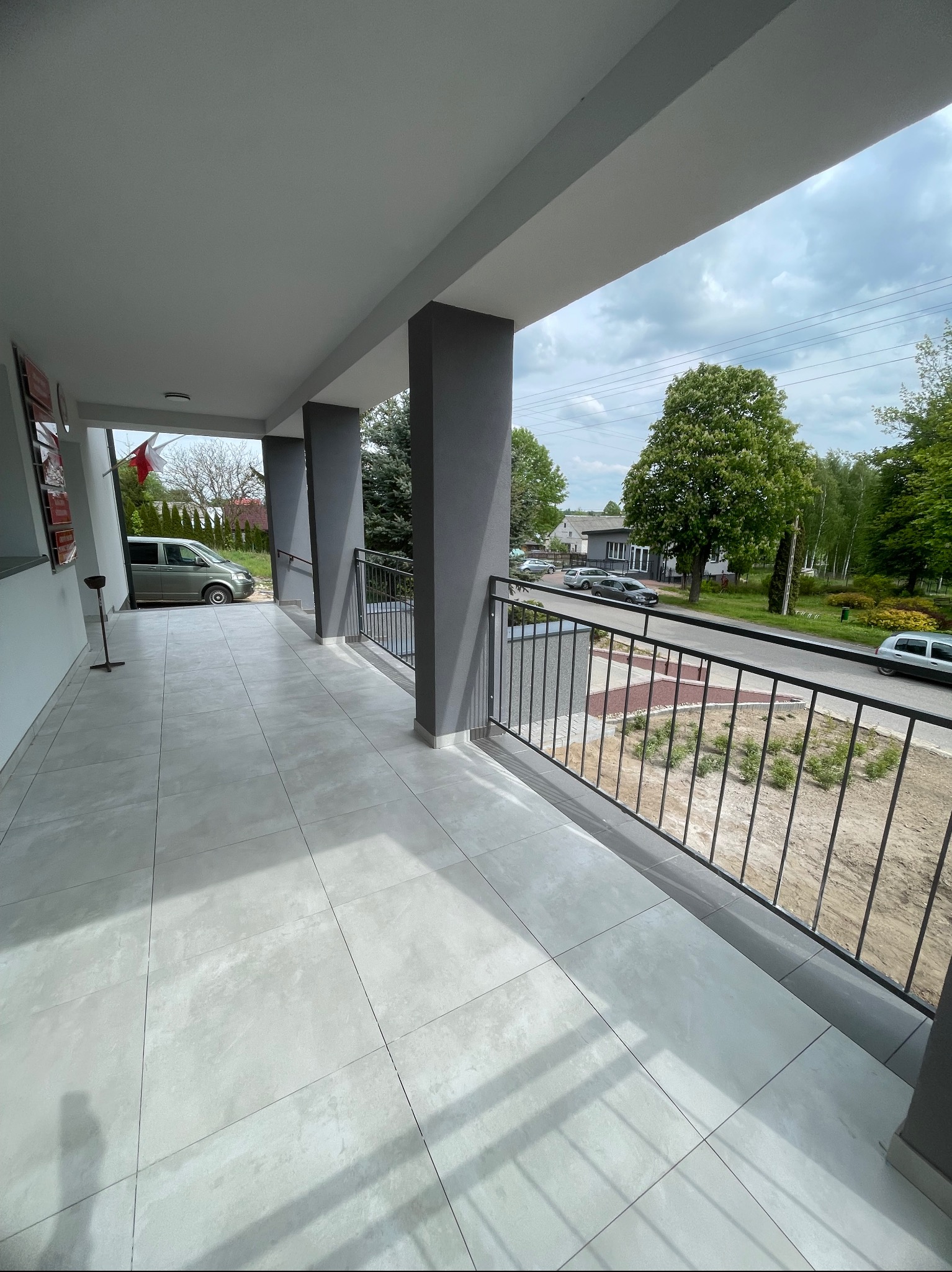 Wykończony balkon z szarymi kolumnami i czarną balustradą. Jasne płytki podłogowe. Widok na zewnątrz z zielenią i zaparkowanymi samochodami.