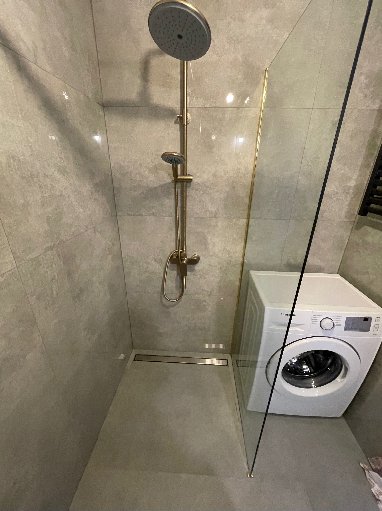 Nowoczesna kabina prysznicowa z deszczownicą w odcieniach szarości, z odpływem liniowym i szklaną ścianką, obok pralka. Minimalistyczny design łazienki.