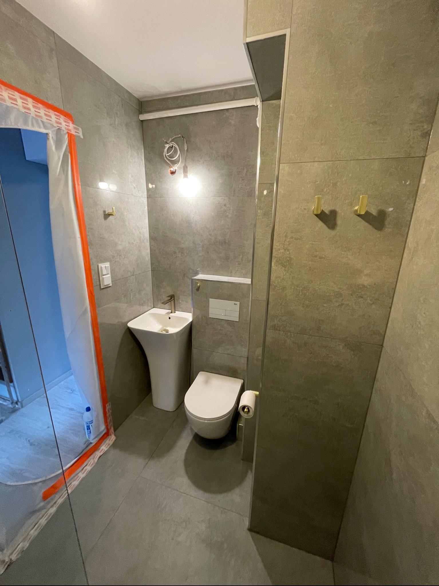 Nowoczesna łazienka w szarościach z wiszącą toaletą, umywalką na postumencie i złotymi haczykami, częściowo zabezpieczona folią ochronną.