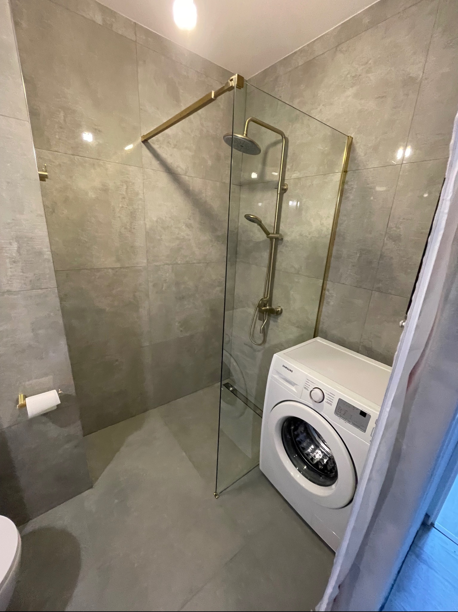 Nowoczesna łazienka z kabiną prysznicową typu walk-in, wykończona szarymi płytkami. Widoczna pralka i złote detale armatury.