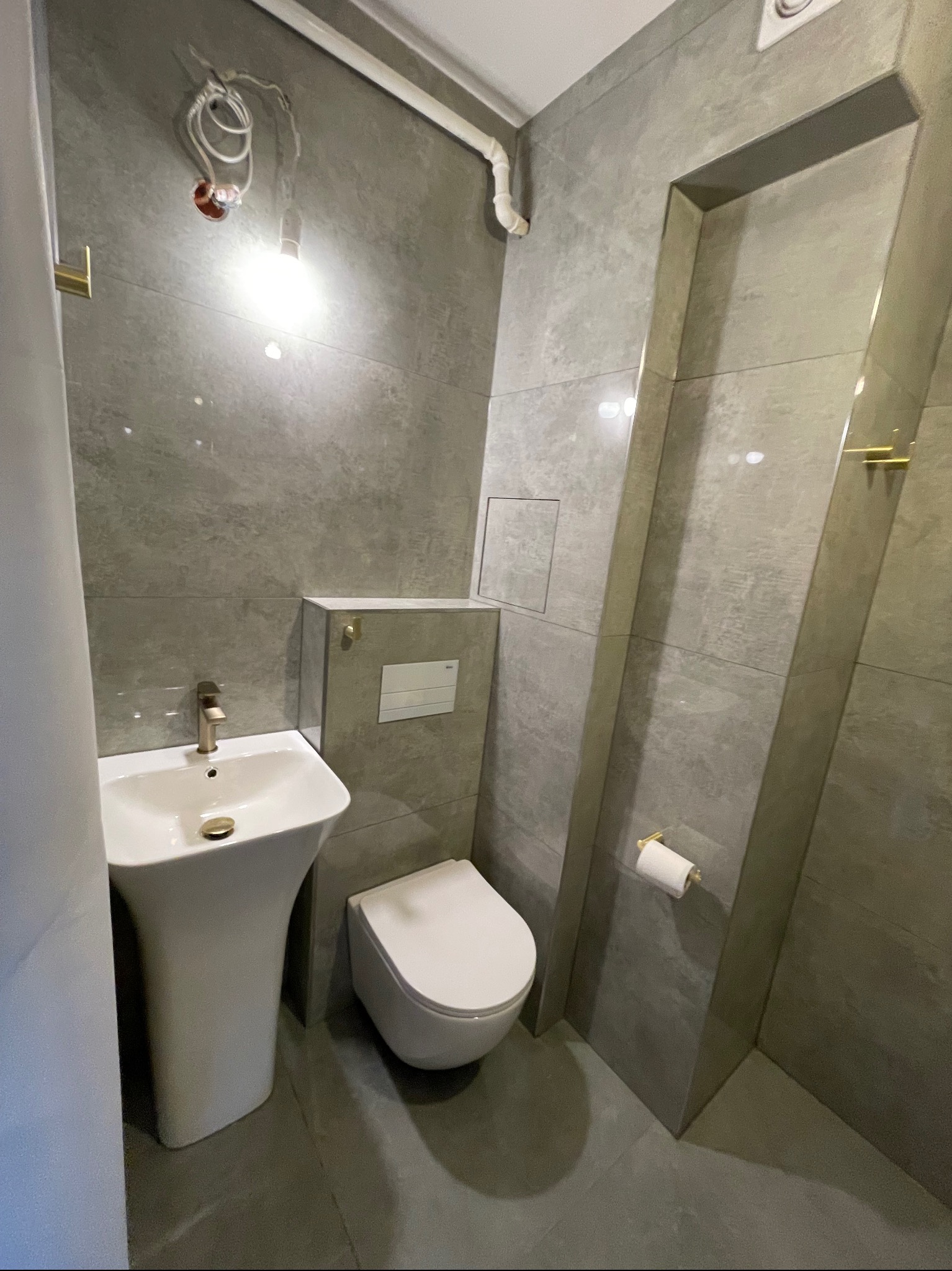 Wykończona łazienka w szarościach: umywalka na postumencie, podwieszana toaleta, płytki imitujące kamień, złote akcenty i surowa żarówka bez oprawy.