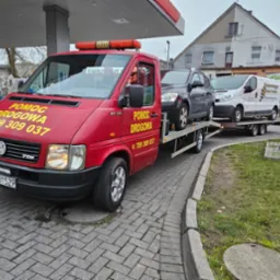 Czerwona laweta Volkswagen LT35 z napisem 'POMOC DROGOWA' i numerem telefonu, przewożąca szare kombi na platformie i biały van na przyczepie, zaparkowana na stacji benzynowej.