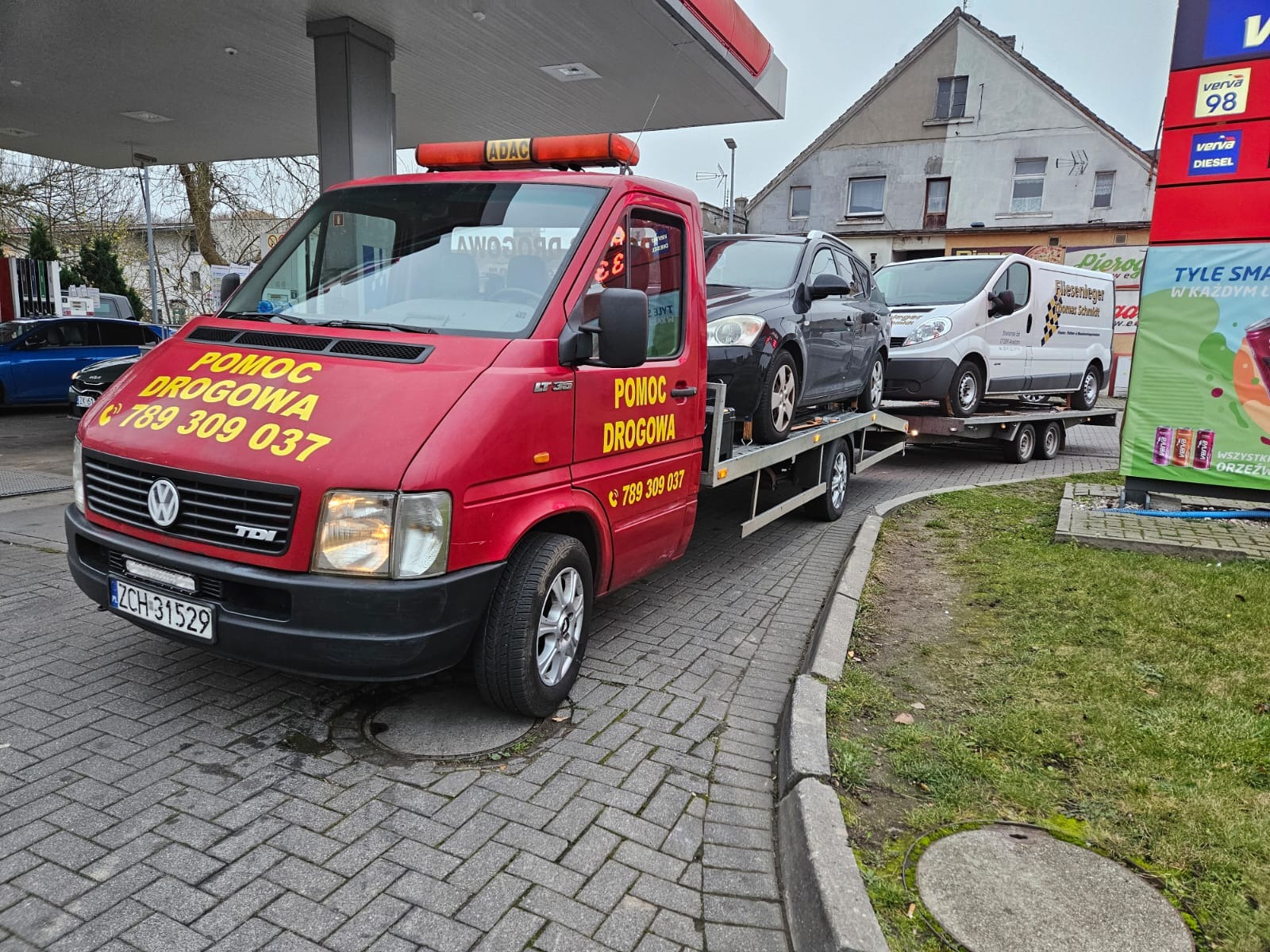 Czerwona laweta Volkswagen LT35 z napisem 'POMOC DROGOWA' i numerem telefonu, przewożąca szare kombi na platformie i biały van na przyczepie, zaparkowana na stacji benzynowej.