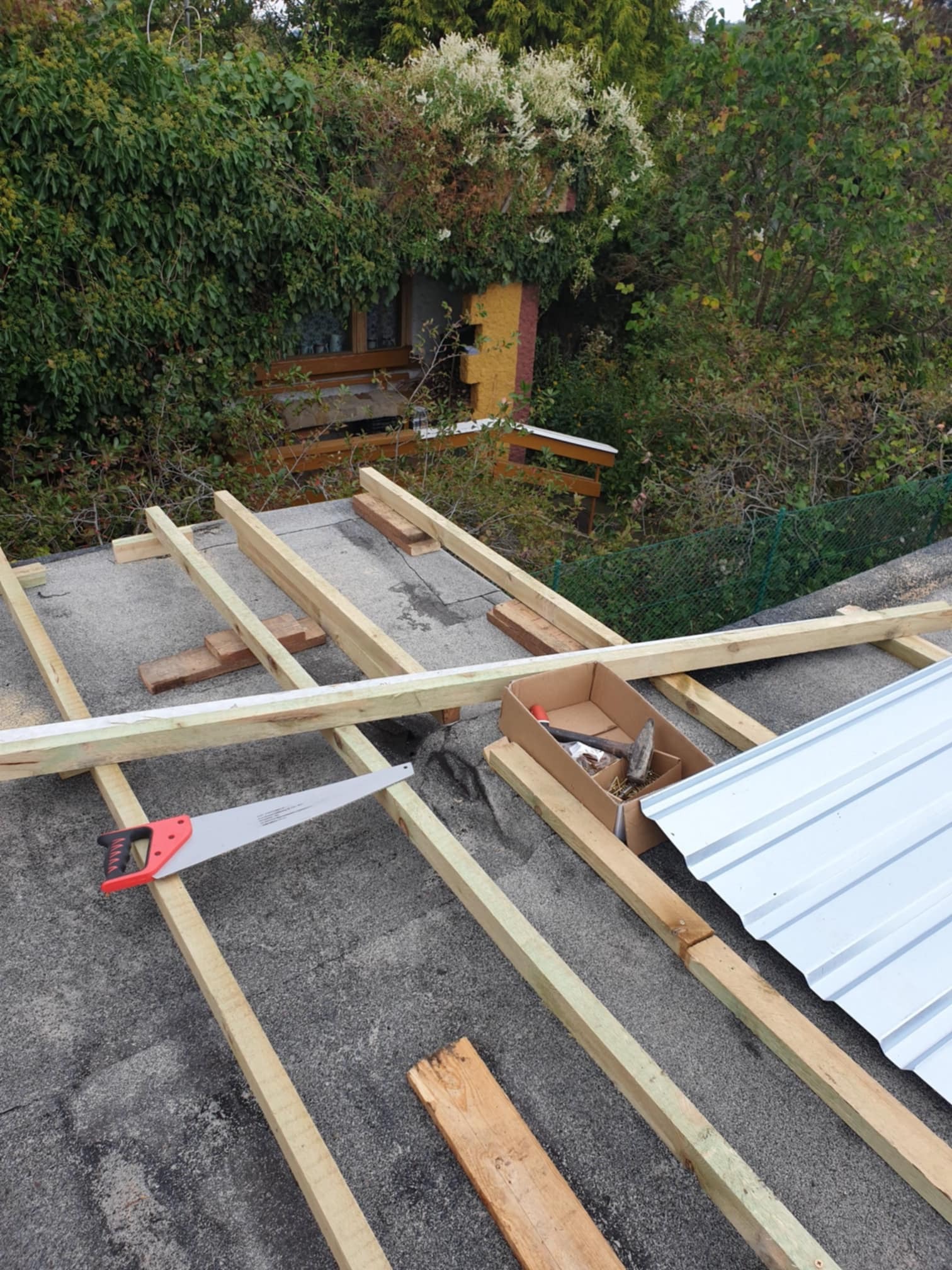 Konstrukcja drewniana z łatami i blachą trapezową na dachu, widoczna piła ręczna i karton z narzędziami, w tle zieleń i budynek.