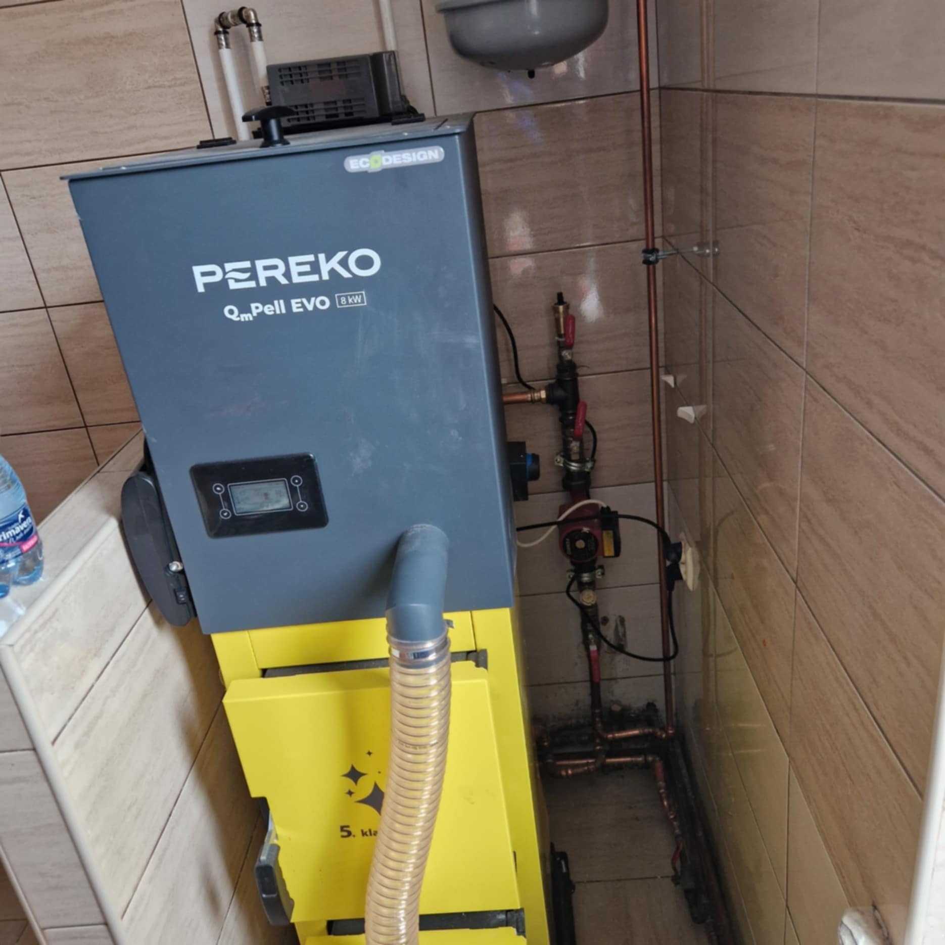 Szary i żółty piec na pellet marki PEREKO Qm Pell EVO 8kW w narożniku wykafelkowanej łazienki, widoczne rury instalacji grzewczej.
