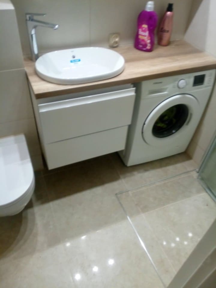 Nowoczesna łazienka z umywalką nablatową, szafką z szufladami, pralką pod blatem i beżowymi płytkami podłogowymi. Widoczne kosmetyki na blacie obok pralki.