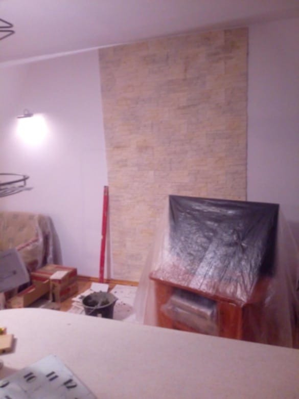 Fragment ściany wyłożony dekoracyjną okładziną kamienną w trakcie montażu, widoczne narzędzia i zabezpieczone meble.