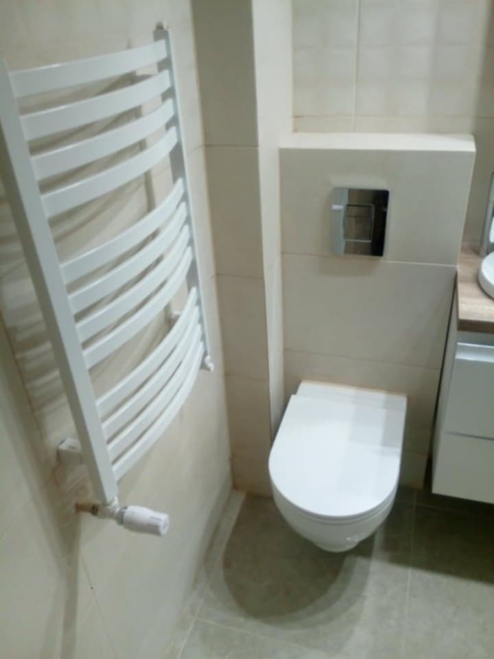 Nowoczesna toaleta podwieszana z chromowanym przyciskiem spłukującym, biały grzejnik drabinkowy z termostatem, beżowe płytki ceramiczne na ścianach i podłodze.