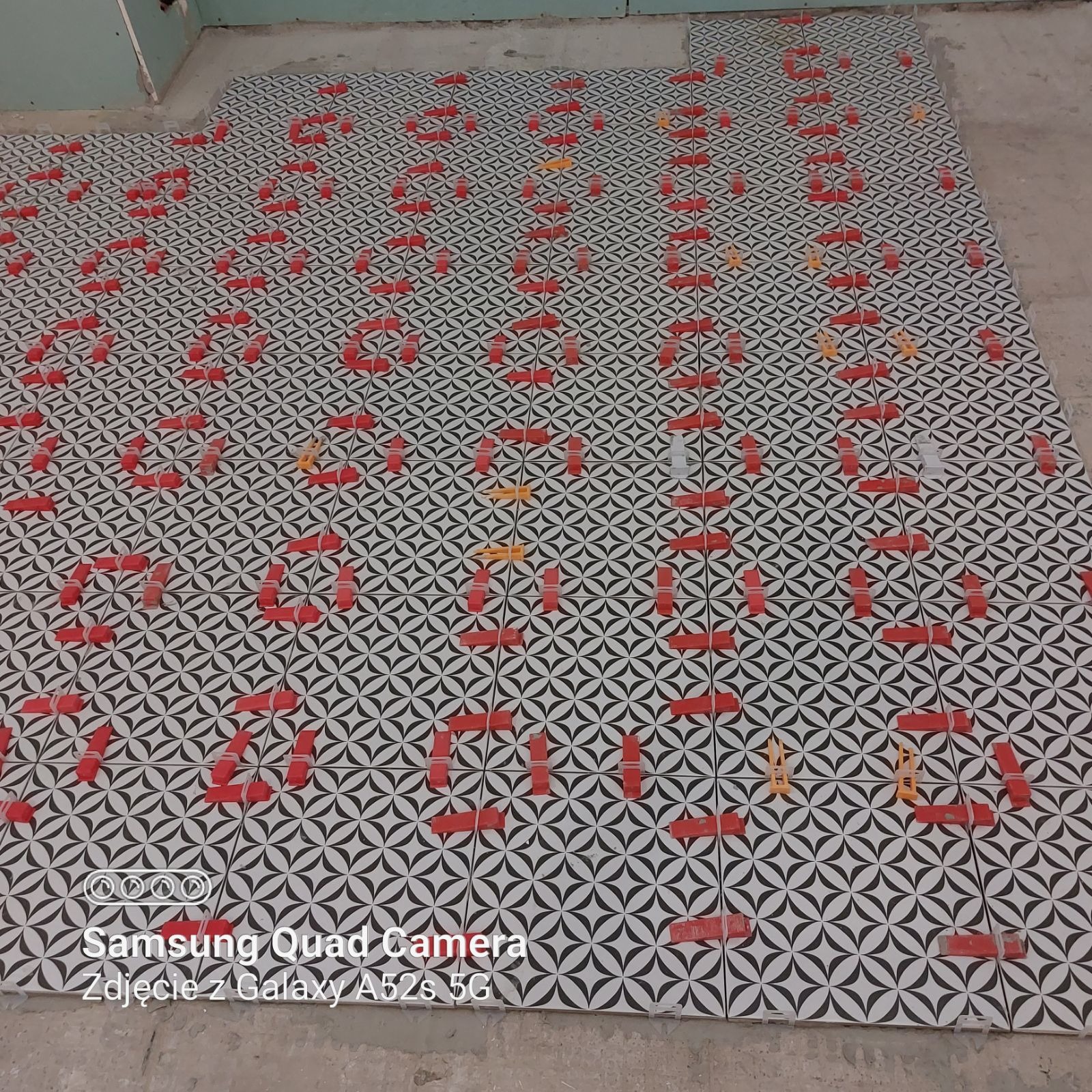 Układanie wzorzystej, biało-czarnej podłogi z użyciem czerwonych i żółtych klinów poziomujących, widoczne fragmenty surowej ściany w tle.