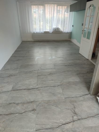 Salon po remoncie z szarymi płytkami podłogowymi imitującymi marmur, widoczne okno z białymi firankami i częściowo pomalowane ściany na kolor miętowy.