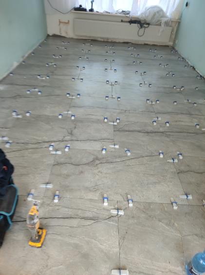 Szare płytki podłogowe ułożone z użyciem systemów poziomowania, widoczna wiertarka akumulatorowa i butelka wody, stanowiące wyposażenie glazurnika.