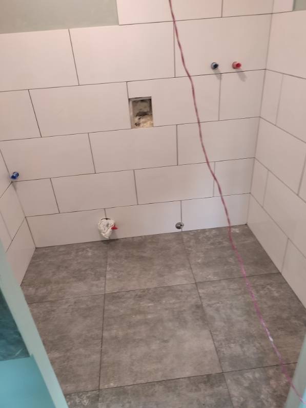 Świeżo ułożone, białe, prostokątne płytki na ścianach i szare, kwadratowe płytki podłogowe w łazience w trakcie remontu, widoczne wystające kable i niezabezpieczone rury.
