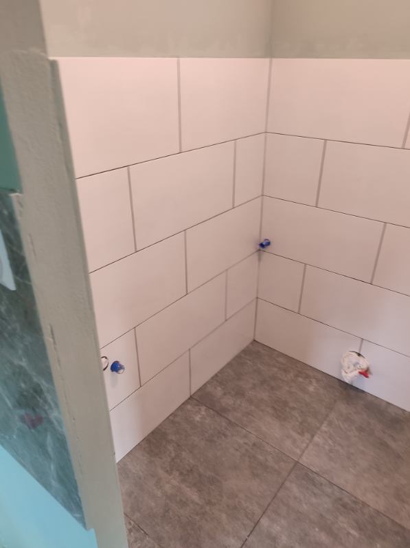 Świeżo ułożone białe płytki ścienne i szare płytki podłogowe w rogu łazienki, z zabezpieczonymi niebieską taśmą rurami wystającymi ze ściany i zakrytą rurą podłączeniową na podłodze, widok z góry.