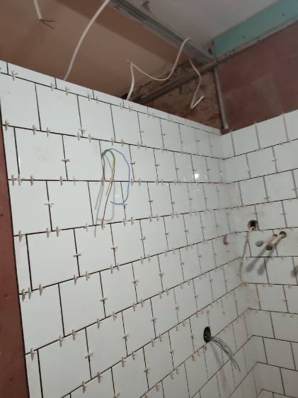 Białe płytki ścienne w trakcie układania w łazience, widoczne krzyżyki dystansowe, wystające przewody elektryczne i rury instalacyjne, surowy stan wykończenia.