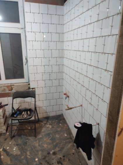 Ściany łazienki w trakcie układania białych, kwadratowych płytek; widoczne krzyżyki dystansowe, rurka instalacji wodnej, okno oraz krzesło z narzędziami w tle.